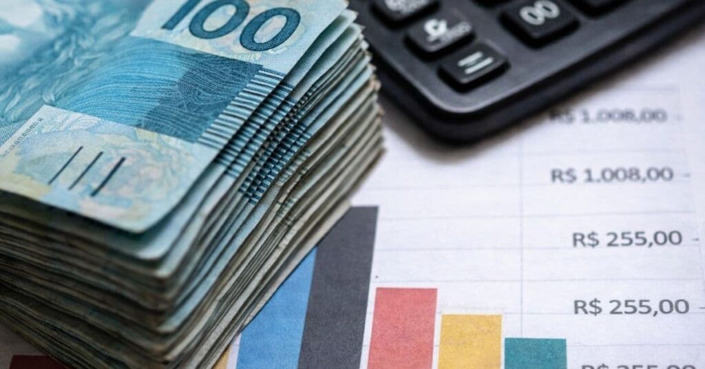 Pilha de cédulas de R$ 100 ao lado de calculadora e gráfico orçamentário, simbolizando o custo dos juros da dívida pública no Brasil.